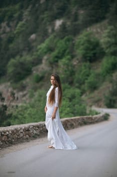 A woman in a white dress is standing on a road. The road is surrounded by trees and rocks. The woman is alone and is looking off into the distance. The scene has a peaceful and serene mood