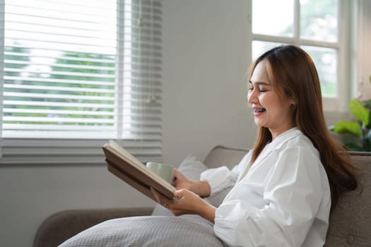 A woman enjoys her morning routine, reading a book and sipping coffee in a cozy home setting by the window.