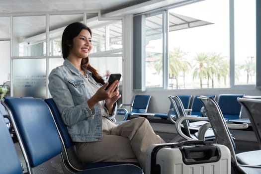 A woman sits in an airport terminal, smiling and using her smartphone while waiting to board her flight. She has a suitcase beside her, ready for her travel adventure.