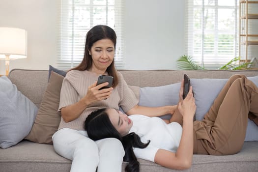 An elderly mother and her adult daughter spend quality time together on a comfortable couch, both engaged with their smartphones in a cozy living room setting.