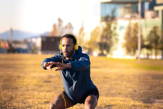 Concentrated young man with sports headphones doing squats. Athlete boy doing squat exercises outdoors.