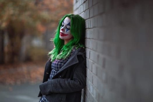 Portrait of a greenhaired girl in chekered dress with joker makeup on a brick wall blurred background.