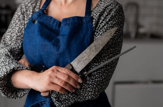Woman Holds Knife And Sharpener In Kitchen Apron, Close-Up Of Hand In Focus