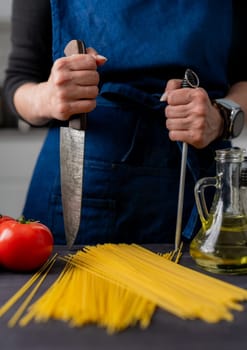 Large Kitchen Knife And Sharpener In Female Hands Are In Close-Up Over Table With Spaghetti, Tomatoes, Spices, And Basil
