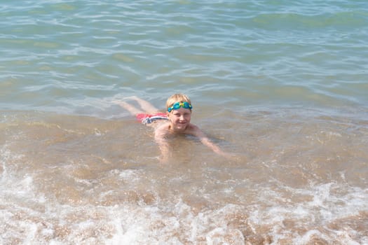 boy 8 years old in swimming goggles lies in the surf on the sandy beach of the blue sea, loves to swim during the summer holidays in Turkey, refreshing swim on a hot sunny day