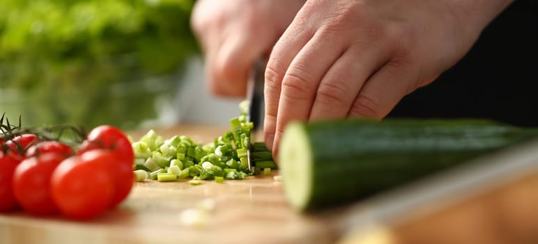 Cook holds knife in hand and cuts on cutting board green onions for salad or fresh vegetable soup with vitamins. Raw food and vegetarian recipe book in modern society popular concept.