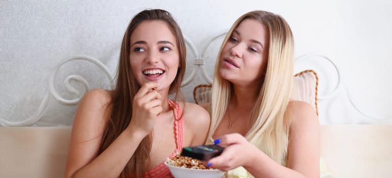Two happy smiling girlfriends eat popcorn in bed watching TV show early morning holding remote control in hand portrait. Joy cozy tenderness lifestyle football match happiness or tv series concept