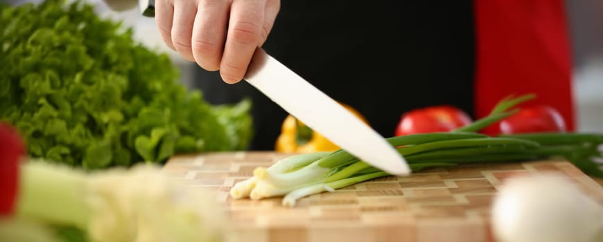 Cook holds knife in hand and cuts on cutting board green onions for salad or fresh vegetable soup with vitamins. Raw food and vegetarian recipe book in modern society popular concept.