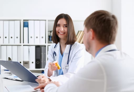 Female medicine doctor write prescription at worktable while patient hand hold jar of pills. Panacea and life save prescribing treatment legal drug store concept. Empty form ready to be used