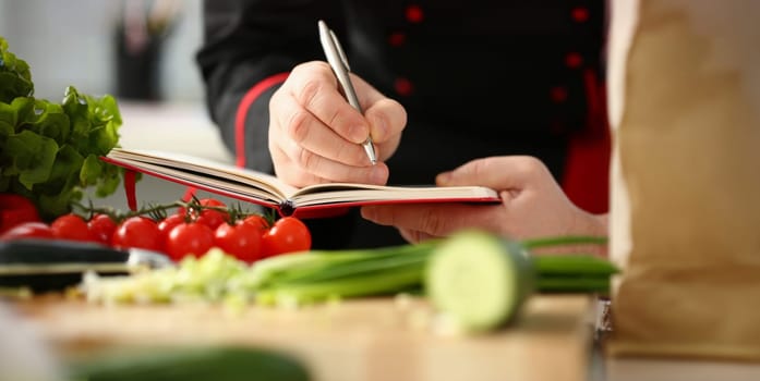 Male cook hand hold siver pen and wright open book of recipes on background of vegetables. Chef writes new combination of taste in home magazine shopping organizer concept list