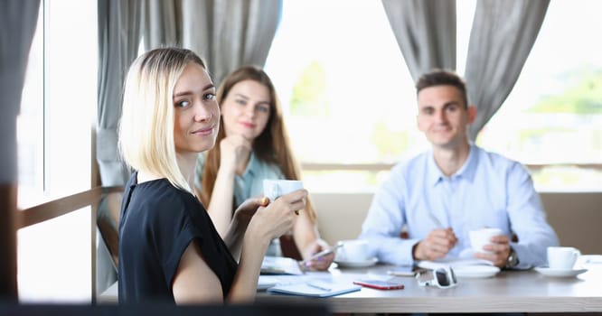 Beautiful smiling cheerful girl at workplace look at camera with colleagues group in background. White collar worker at cafe modern lifestyle client visit profession train concept coffe drink