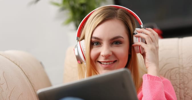 young beautiful blond woman sit on the sofa in livingroom hold tablet in arms listen to music sunny morning concept