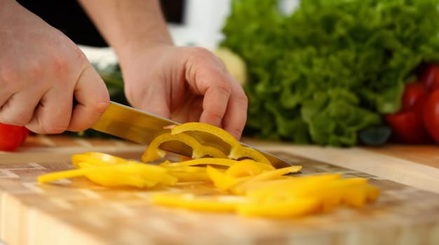 Cook holds knife in hand and cuts on cutting board yellow pepper for salad or fresh vegetable soup with vitamins. Raw food and vegetarian recipe book in modern society popular concept.