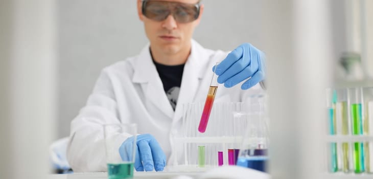 A male chemist holds test tube of glass in his hand overflows a liquid solution of potassium permanganate conducts an analysis reaction takes various versions of reagents using chemical manufacturing.
