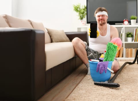 A young smiling man holds a smartphone in his hand demonstrating it to the camera advertising a mobile application to search for cleaning