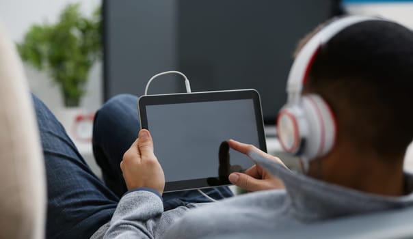Male hand holds tablet pad in home setting while sitting on the couch engaged an internet surfing using application to press a finger on the display leisure listering music concept closeup.