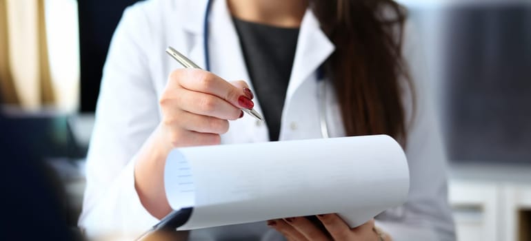 Female doctor hand hold silver pen filling patient history list at clipboard pad. Physical exam er disease prevention ward round visit check 911 prescribe remedy healthy lifestyle concept