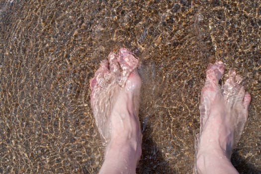 Female feet barefoot in waves of foamy water on a sandy beach on a summer day, top view of female feet, woman on the beach getting her feet wet in warm sea water, carefree vacation by the sea,