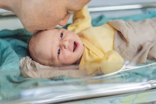 Mom kisses her newborn baby who is resting in a transparent cradle in the hospital. This tender moment emphasizes the bond between mother and child, creating a cozy and safe atmosphere for the infant