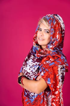 A woman wearing a red, blue and white scarf stands in front of a pink background. She is smiling and she is happy