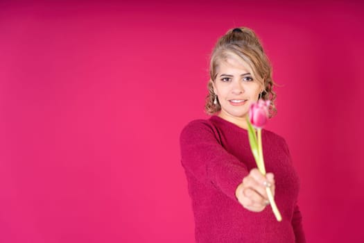 Young pretty woman smiling looking at camera offering a tulip flower, wearing a red t-shirt on a red background with copy space. Gifts concept.