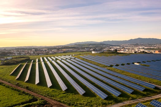 Aerial view field with rows of solar panels, set against a backdrop of sky, clouds, and natural landscapes. Green grass contrasts with the infrastructure. Mountain on background and beautiful sunset