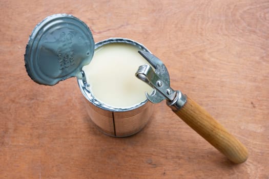 Opened metal can of condensed milk and metal can opener on brown wooden background.