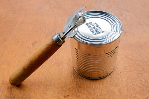 Unopened metal can and can opener on brown wooden background.