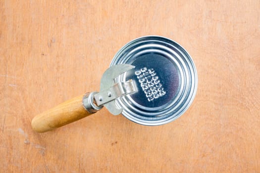 Unopened metal can and can opener on wooden brown background, top view.