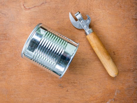 White metal can and can opener on brown wooden background, top view. Concept of food on a camping trip.