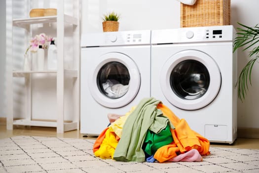 A pile of clean, colorful laundry sits on the floor in front of two white washing machines in a modern laundry room.