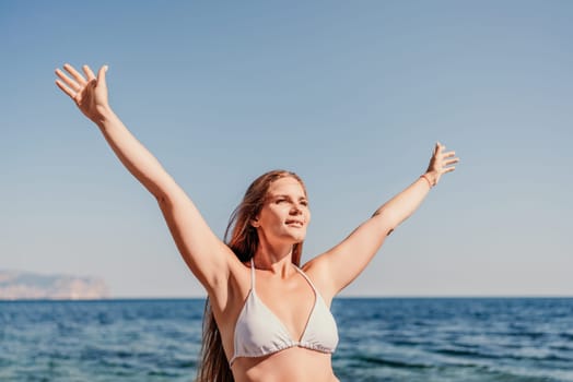 A woman in a bikini is standing on the beach, with her arms raised in the air. Concept of freedom and joy, as the woman is enjoying her time at the beach