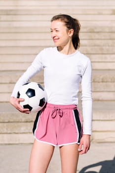 smiling sports woman in a concrete soccer court with a ball under her arm, concept of football lifestyle and urban sport in the city