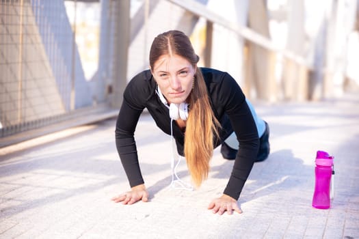 Sporty young woman with sports headphones doing push-ups smiling looking at the camera outdoors.