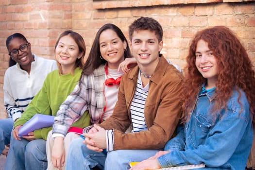 Multiracial group of students sitting looking at the camera laughing outdoors at the university. Young community of people having fun together.