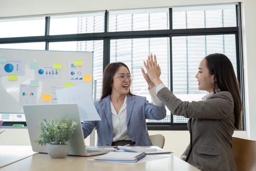 Two young female accountants celebrate their success in a modern office, high-fiving and smiling with charts and graphs in the background.