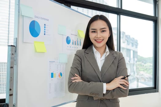 A confident young accountant standing in a modern office, smiling with arms crossed, in front of a whiteboard displaying financial charts and graphs.