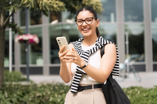 A young professional, phone in hand, walks through an urban park with a confident smile. Dressed stylishly, she balances her roles as a student and businesswoman, symbolizing modern career success.