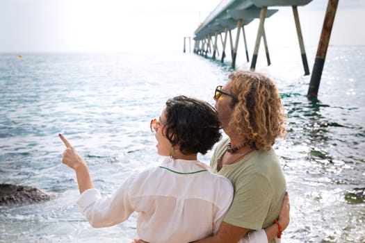 Smiling couple in sunglasses enjoying vacation hugging each other pointing and looking at the sea.