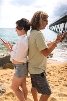 Middle-aged man and woman sitting on the beach with surprised face, browsing telephones apps. Concept:Holidays y Technology