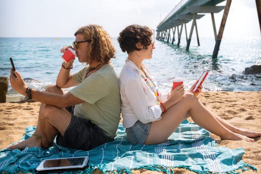 Middle-aged man and woman sitting on the beach with surprised face, browsing telephones apps. Concept:Holidays y Technology