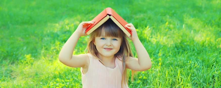 Happy little girl child reading a book sitting on green grass in summer park