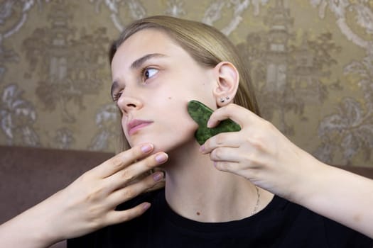 Young girl doing facial massage with gua sha scraper for skin rejuvenation, maintaining freshness and youth of facial skin and body care at home