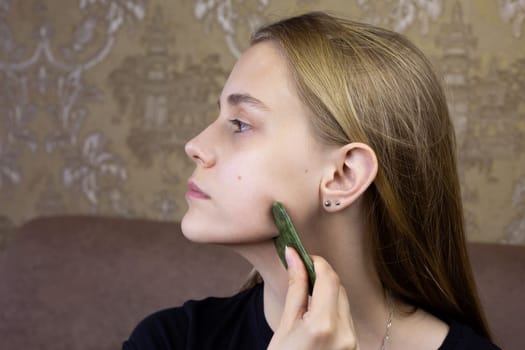 Profile of European girl who does facial massage using gua sha scraper to take care of health of skin and body, procedure for cleaning face and stretching wrinkles using special scraper