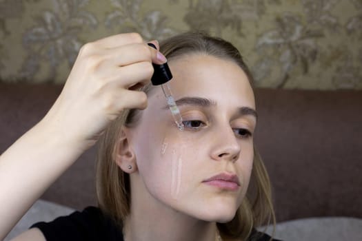 European girl is dripping special massage oil for skin care onto her face, pipette with dosage of product for healthy facial skin