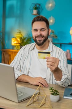 Happy businessman using bank credit card and laptop for online shopping payments at home office. Business transaction. Smiling guy freelancer purchases with netbook E-banking app service. Vertical