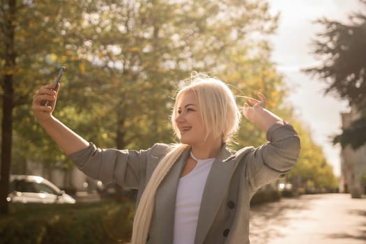 A woman is taking a picture of herself with her cell phone. She is wearing a gray jacket and scarf. The scene is set in a city with trees and cars in the background