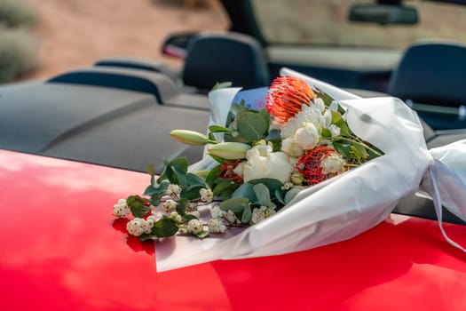A bouquet of flowers is sitting on the hood of a red car. The flowers are white and red, and they are arranged in a vase. Concept of romance and love