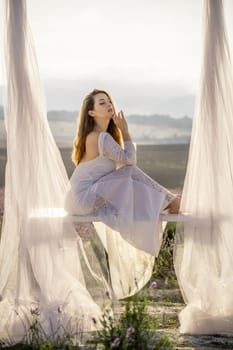 A woman is sitting on a bench under a white canopy. She is wearing a white dress and has long hair. The scene is set in a field with purple flowers