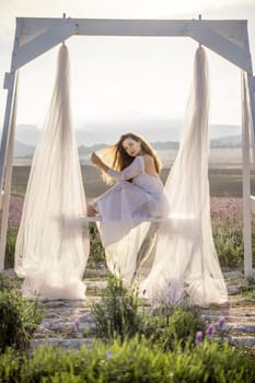 A woman is sitting on a bench under a white canopy. She is wearing a white dress and has long hair. The scene is set in a field with purple flowers